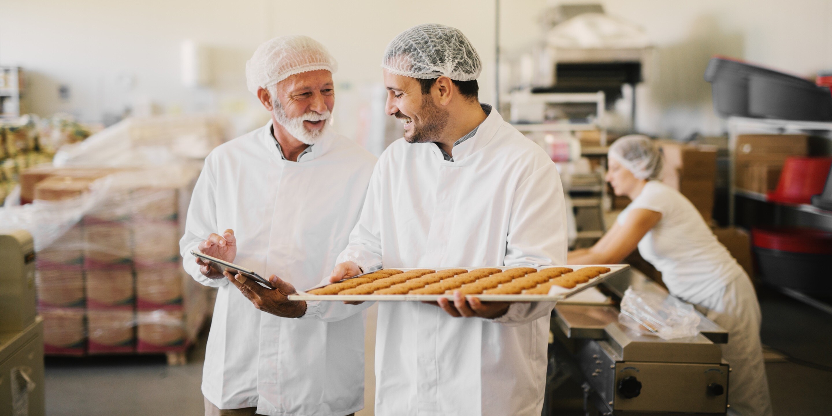 Picture of two employees in sterile clothes in food factory smiling and talking. Younger man is holding tray full of fresh cookies while the older is holding tablet and checking production line.