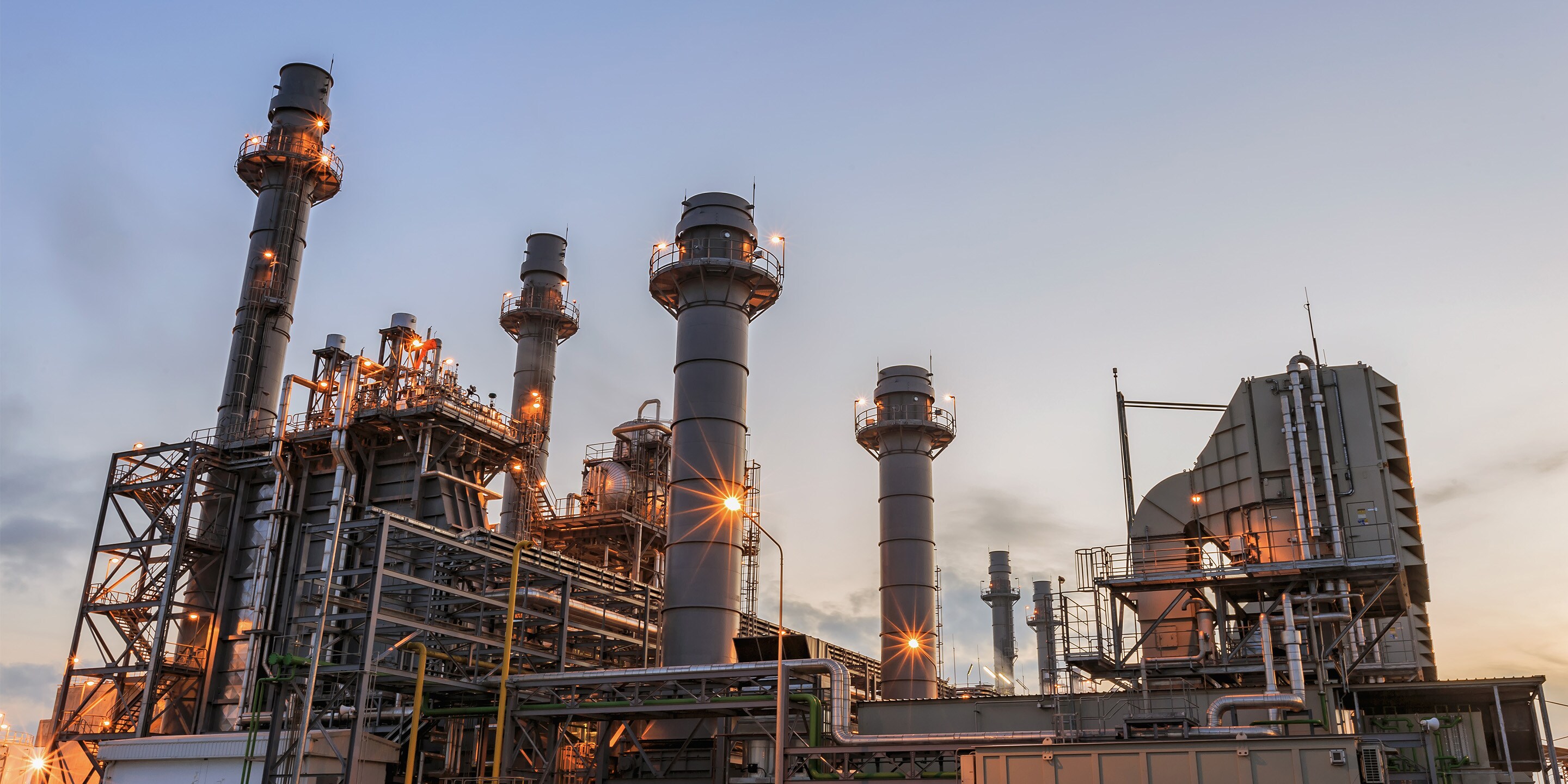 Industrial power plant with tall chimneys and piping, lit at dusk, highlighting energy production infrastructure under a clear sky.